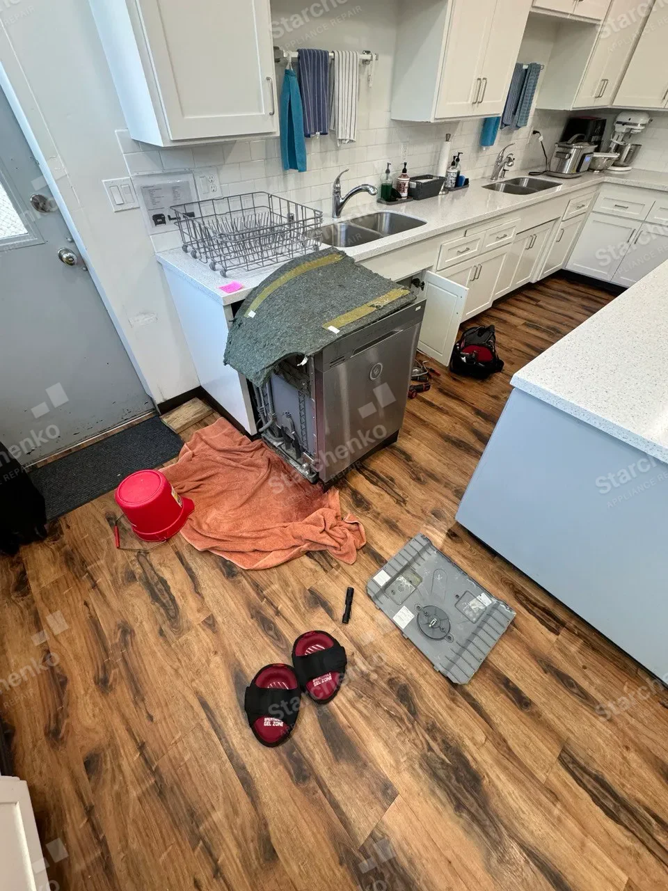 Stainless Amana dishwasher pulled out with its base pan removed and set on the kitchen floor