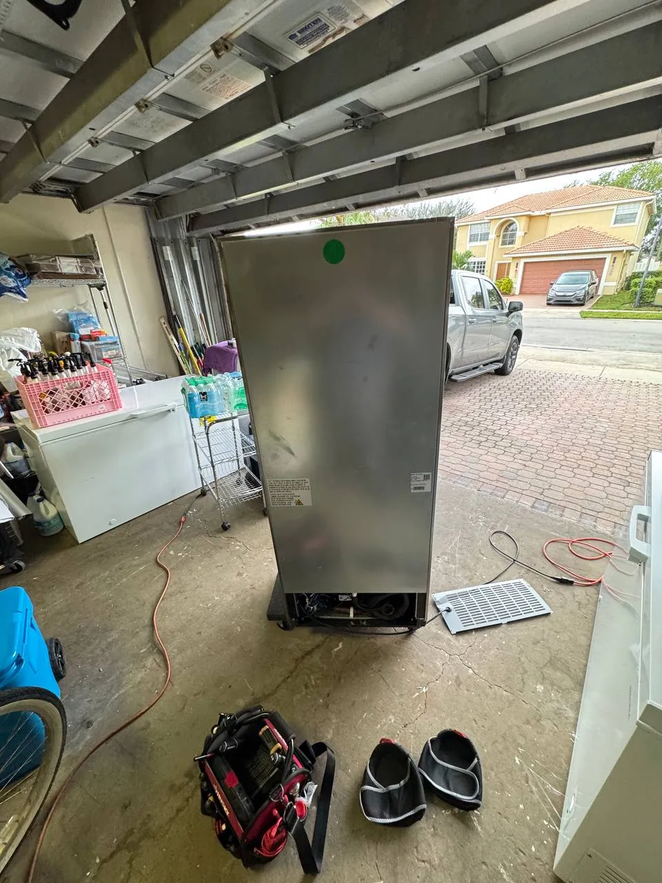Rear of a stainless commercial reach-in cooler in a garage with its lower machine compartment open