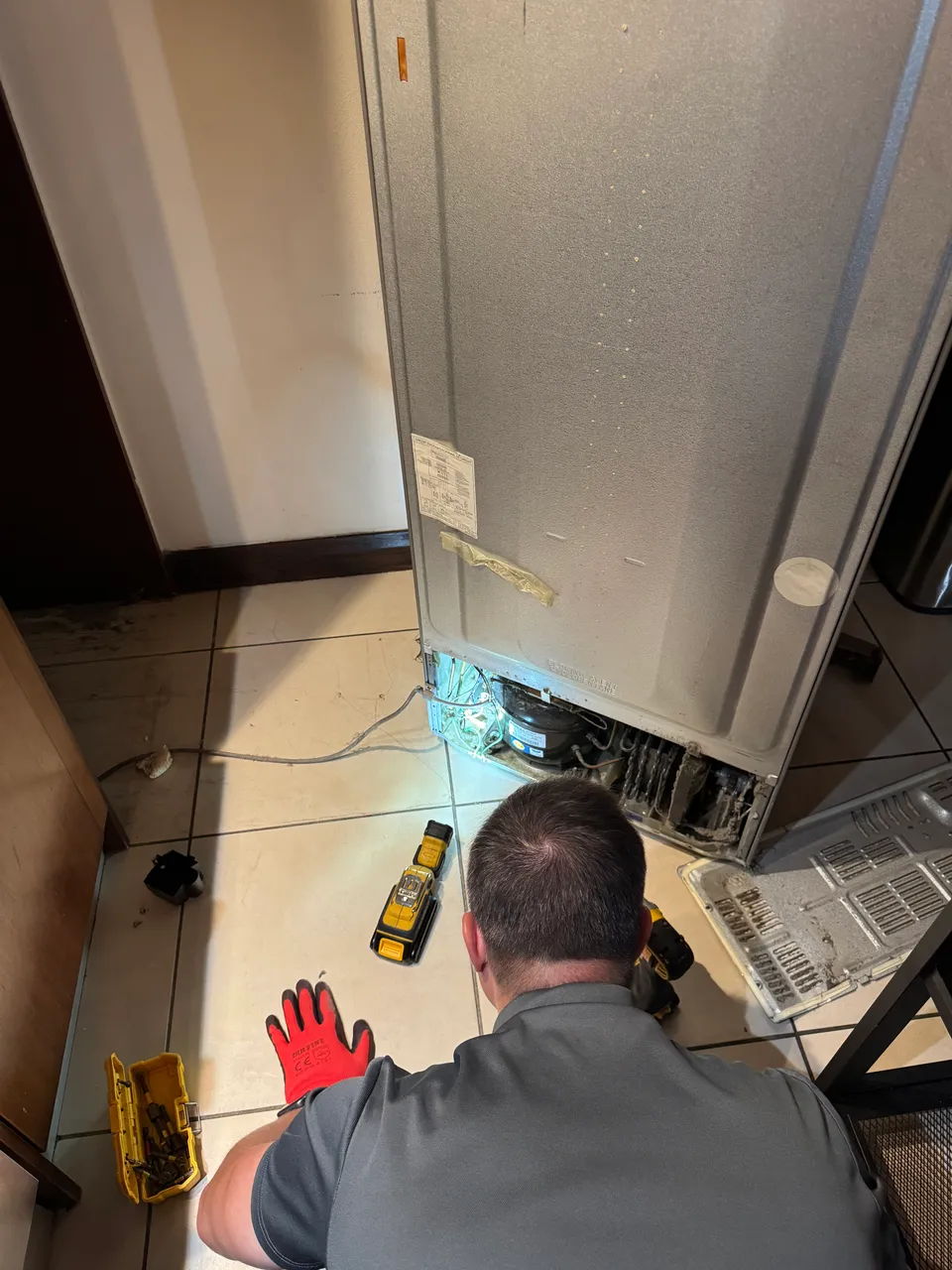 Technician working at the open compressor compartment at the back of a refrigerator, condenser grille removed