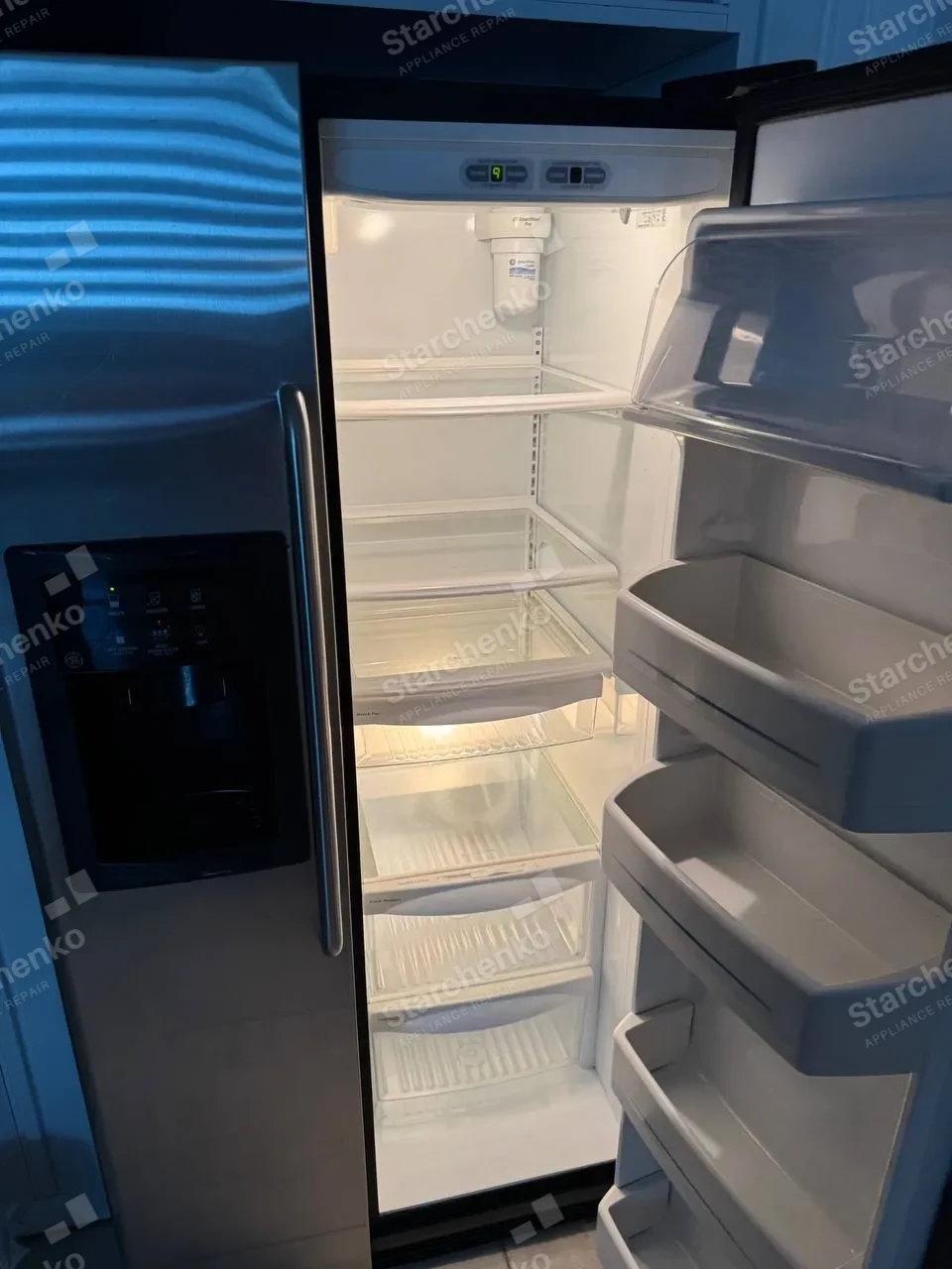 GE side-by-side refrigerator standing open and empty with the water filter in the ceiling of the fresh-food section