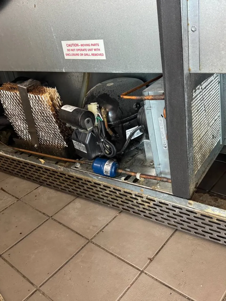 Commercial refrigerator compressor compartment with a clogged condenser coil and blue filter drier above a tile floor