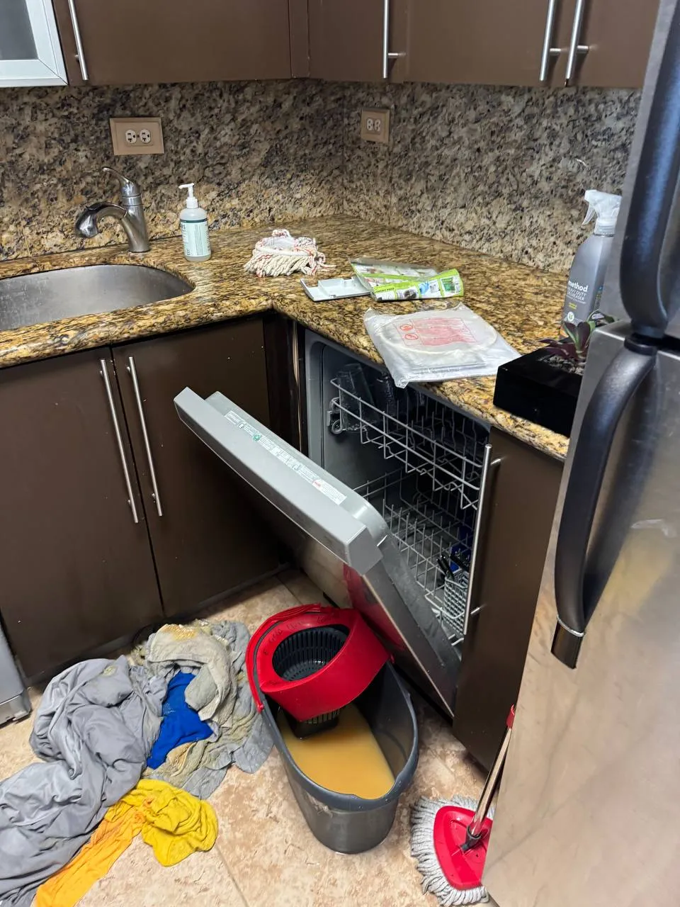 Open Whirlpool dishwasher beneath a granite counter with water collected in a bucket on the floor