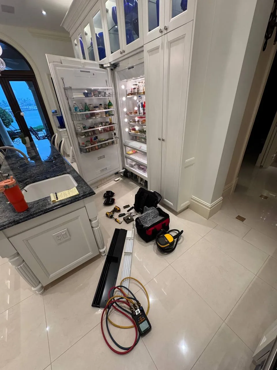Built-in Miele refrigerator with both doors open and the lower grille off, gauges and a tool bag on the floor