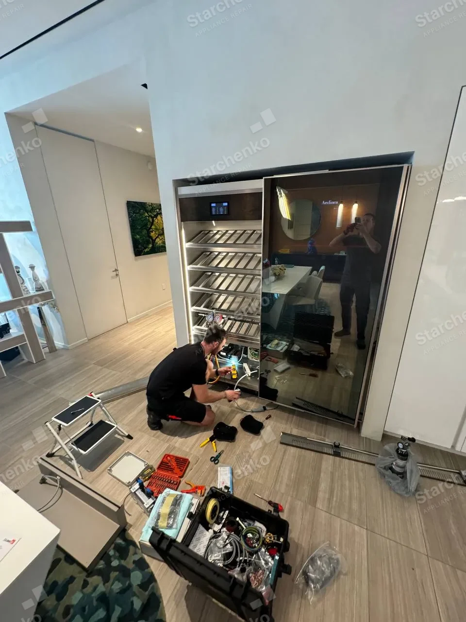 ArteVino technician kneeling at the base of a built-in glass-door wine cooler with refrigerant gauges connected