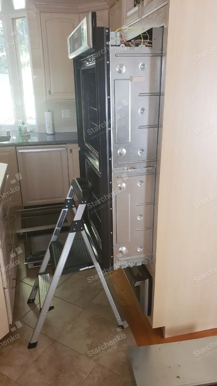 Whirlpool repair: Double wall oven pulled from cabinet with side panels and top wiring exposed beside a ladder.