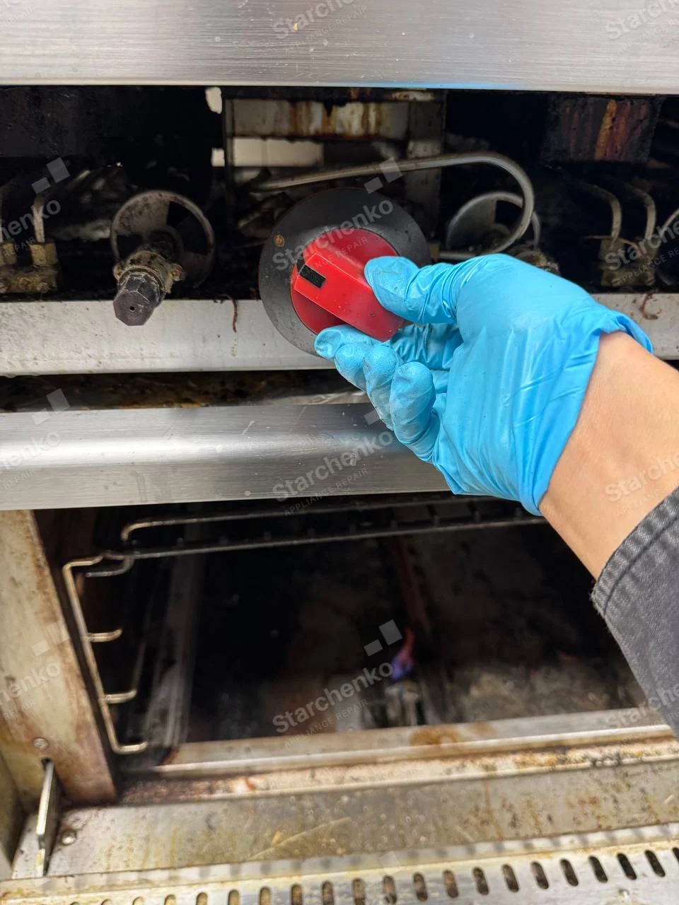 Garland repair: Close-up commercial gas range with oven open and technician adjusting detached red knob at control shaft, no maker mark.
