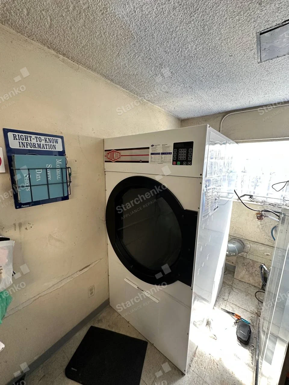 Large Milnor tumble dryer with side service panel open in a laundry room.