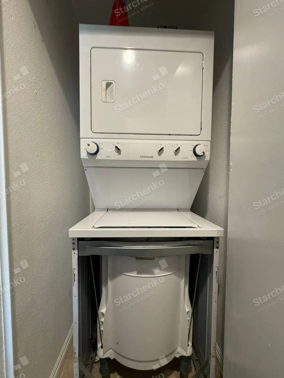 Frigidaire name centered on shared controls, lower washer front panel removed exposing tub below upper dryer.