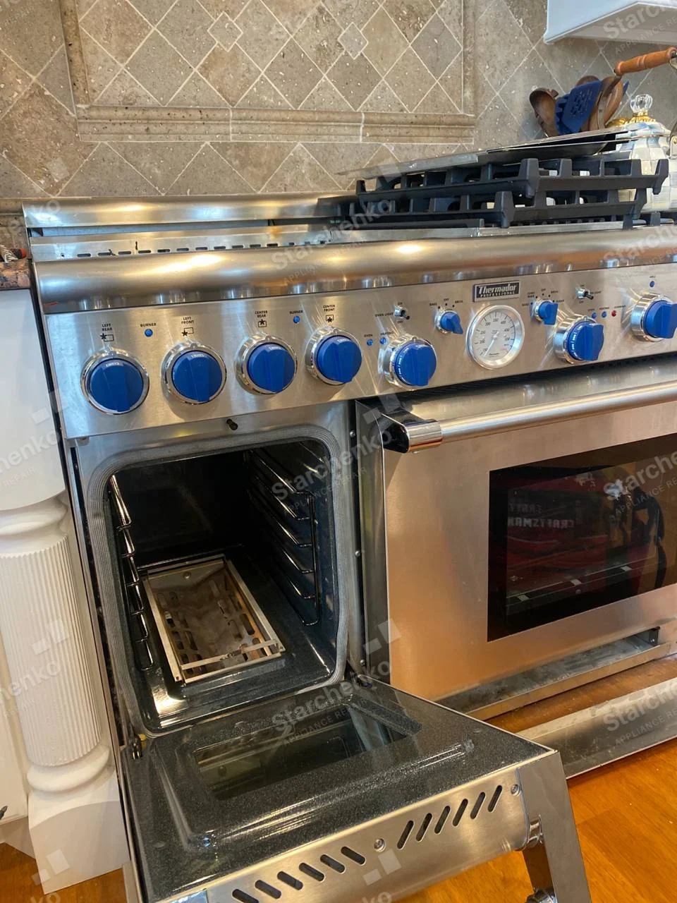 Thermador badge above large oven door, row of blue knobs and smaller secondary oven open at left.