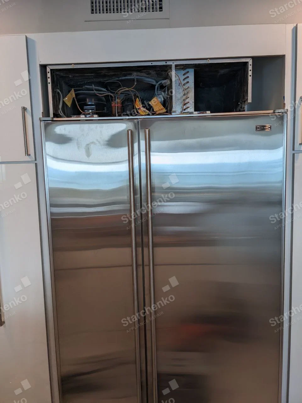 Sub-Zero stainless side-by-side refrigerator with top grille removed and machinery exposed.