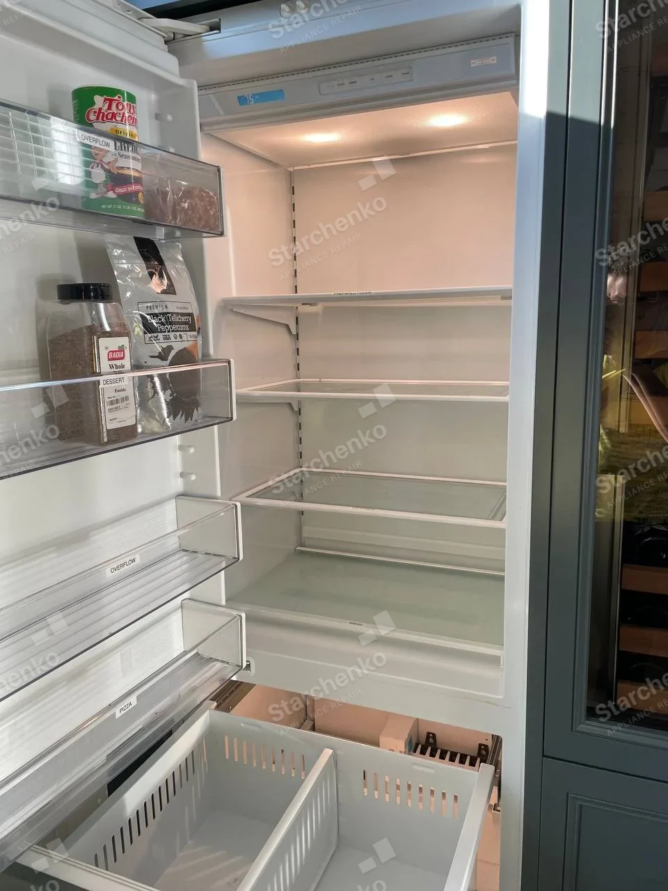 Miele repair: Open built-in refrigerator showing illuminated fresh-food shelves and pull-out freezer baskets.
