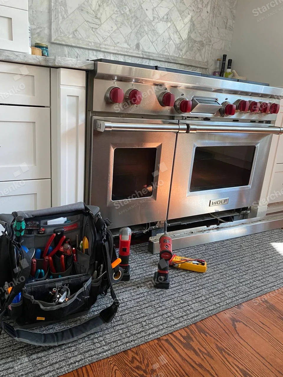 Wolf professional dual-oven gas range with red knobs and lower kick panel removed, tools on floor.