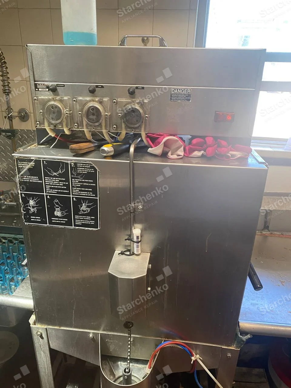Rear service side of a CMA commercial dishwasher with three chemical dosing pumps and wash-arm cleaning instructions.