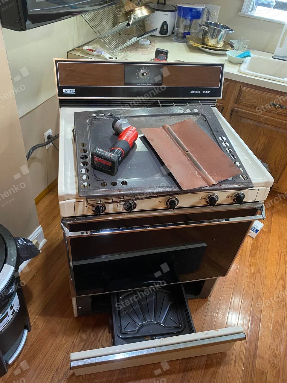 Roper gas range with cooktop panel lifted, drill resting on top and lower drawer pulled out.