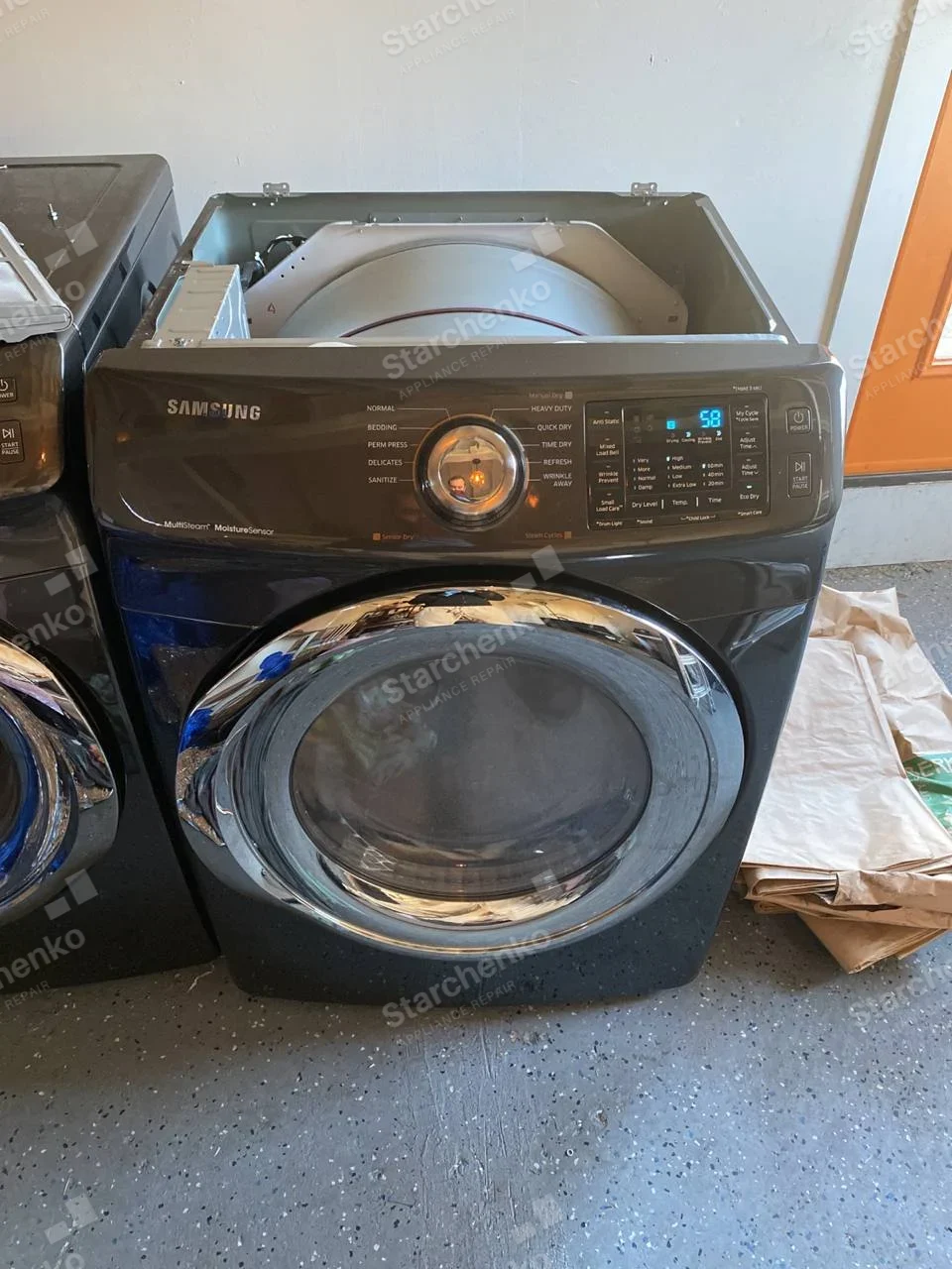 Samsung front-load dryer with top cover removed, metal drum shell visible and display lit.