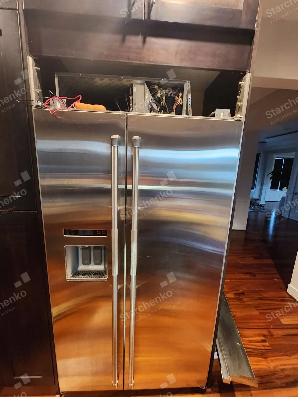 GE Monogram repair: Full-height stainless built-in side-by-side refrigerator with door dispenser, upper mechanical cover removed exposing control wiring.