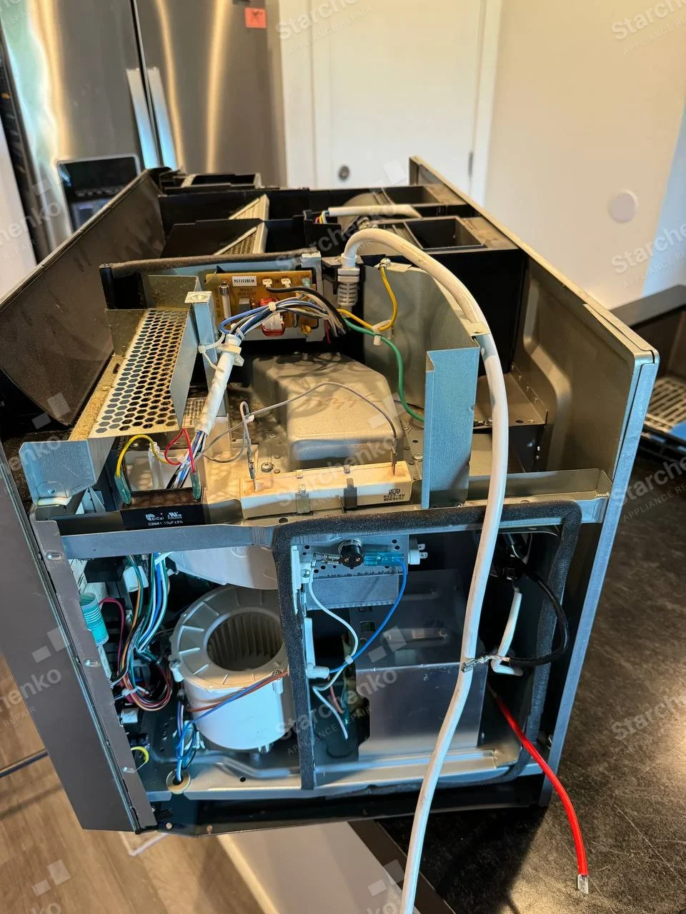 Samsung repair: Removed microwave chassis with outer cover off, high-voltage components and cooling blower exposed, refrigerator in background is separate.
