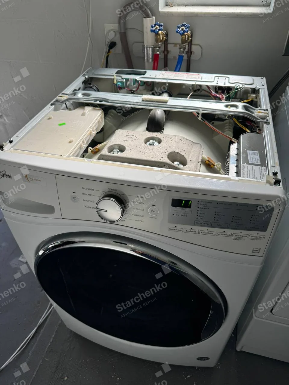 White front-load washer with top removed, exposing tub weights and electronics, Whirlpool lettering on upper-left fascia.