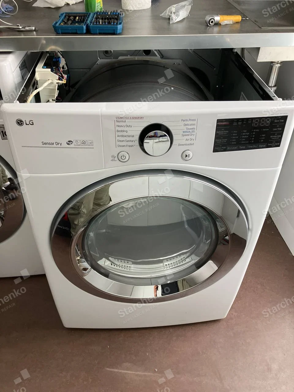 White LG dryer with upper panel removed, exposing drum and electrical module.