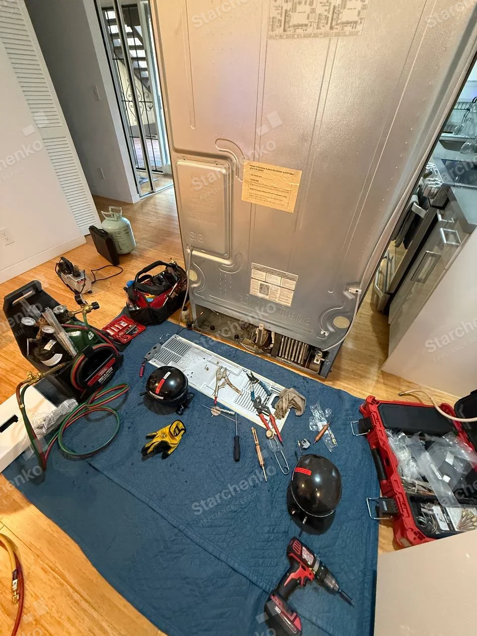 Whirlpool repair: Rear of upright refrigerator exposed, detached compressor and service gauges on floor, no legible refrigerator brand.