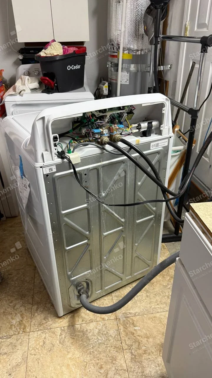 Amana repair: Rear panel of top-load washer open, showing water inlet valves, hoses and control electronics, no maker mark.