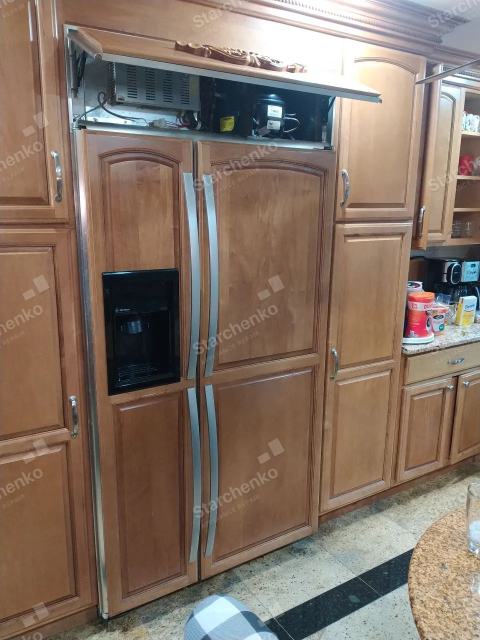 GE Monogram repair: Monogram logo visible on left-door water dispenser of paneled built-in side-by-side refrigerator, top machinery grille open.