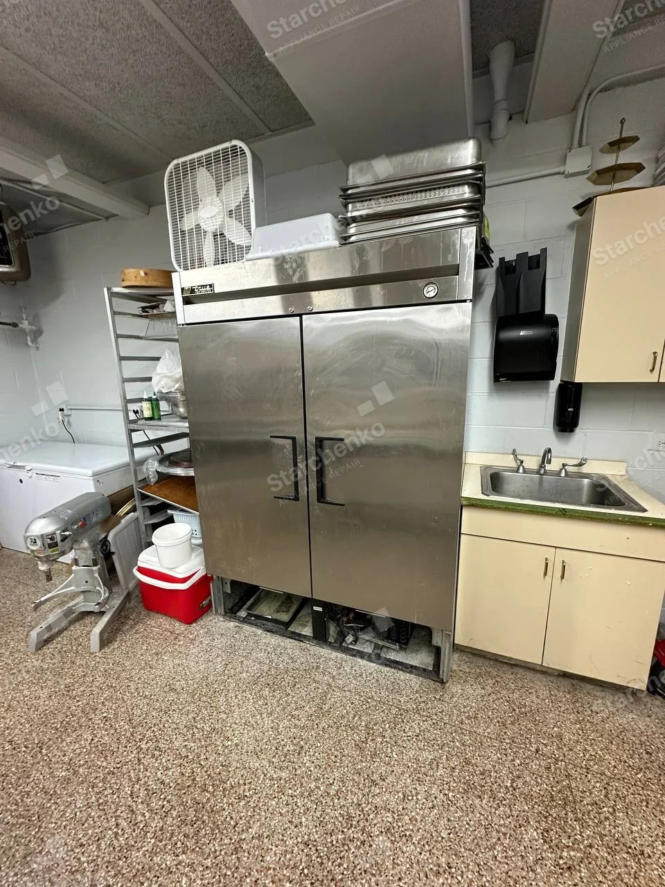Arctic Air repair: Stainless two-door upright commercial refrigerator with lower mechanical access panel removed, neighboring mixer not main appliance.