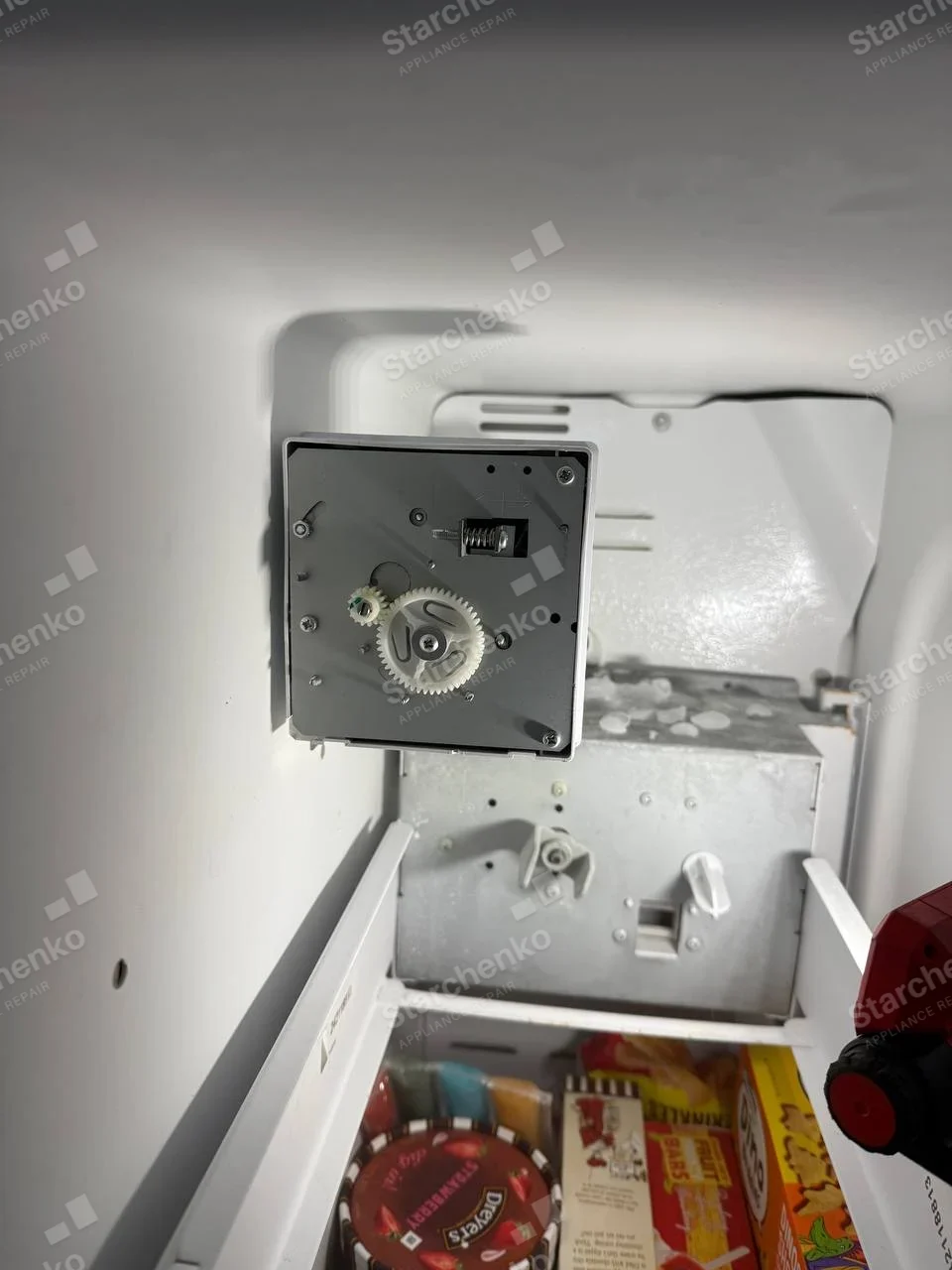 GE repair: Ice-maker gear and motor removed inside a household freezer compartment, main refrigerator brand not visible.
