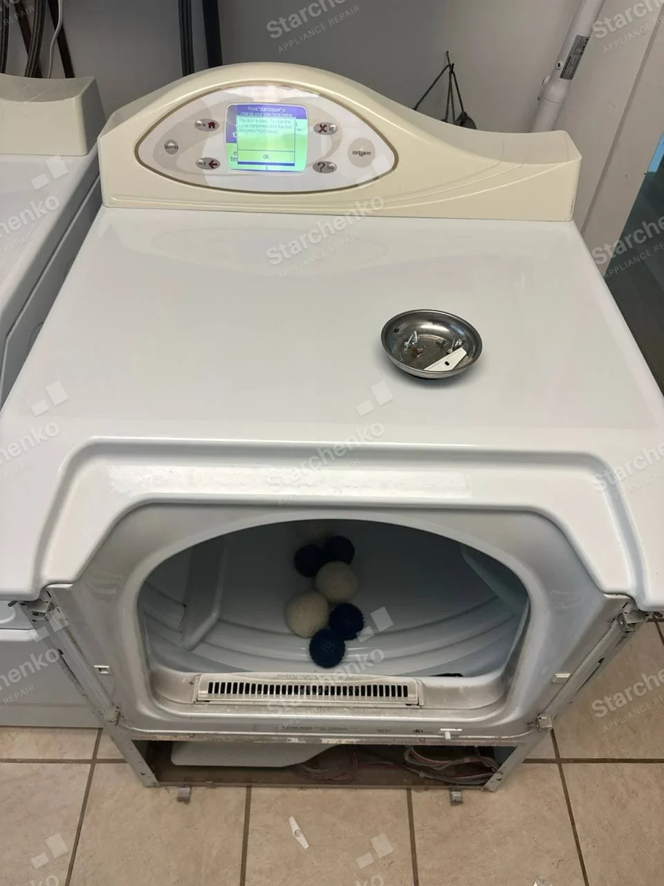 Electrolux repair: Front-load dryer with front and lower panel removed, lint balls and small parts placed atop casing, display lit.