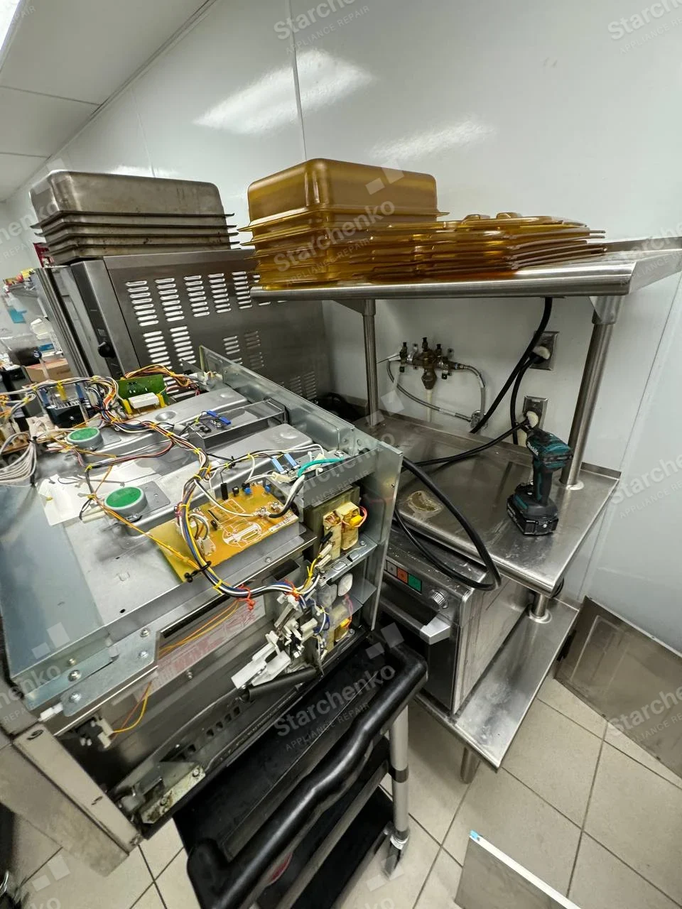 Amana repair: Metal microwave chassis in foreground with top cover removed, control board and wiring exposed, commercial kitchen setting.