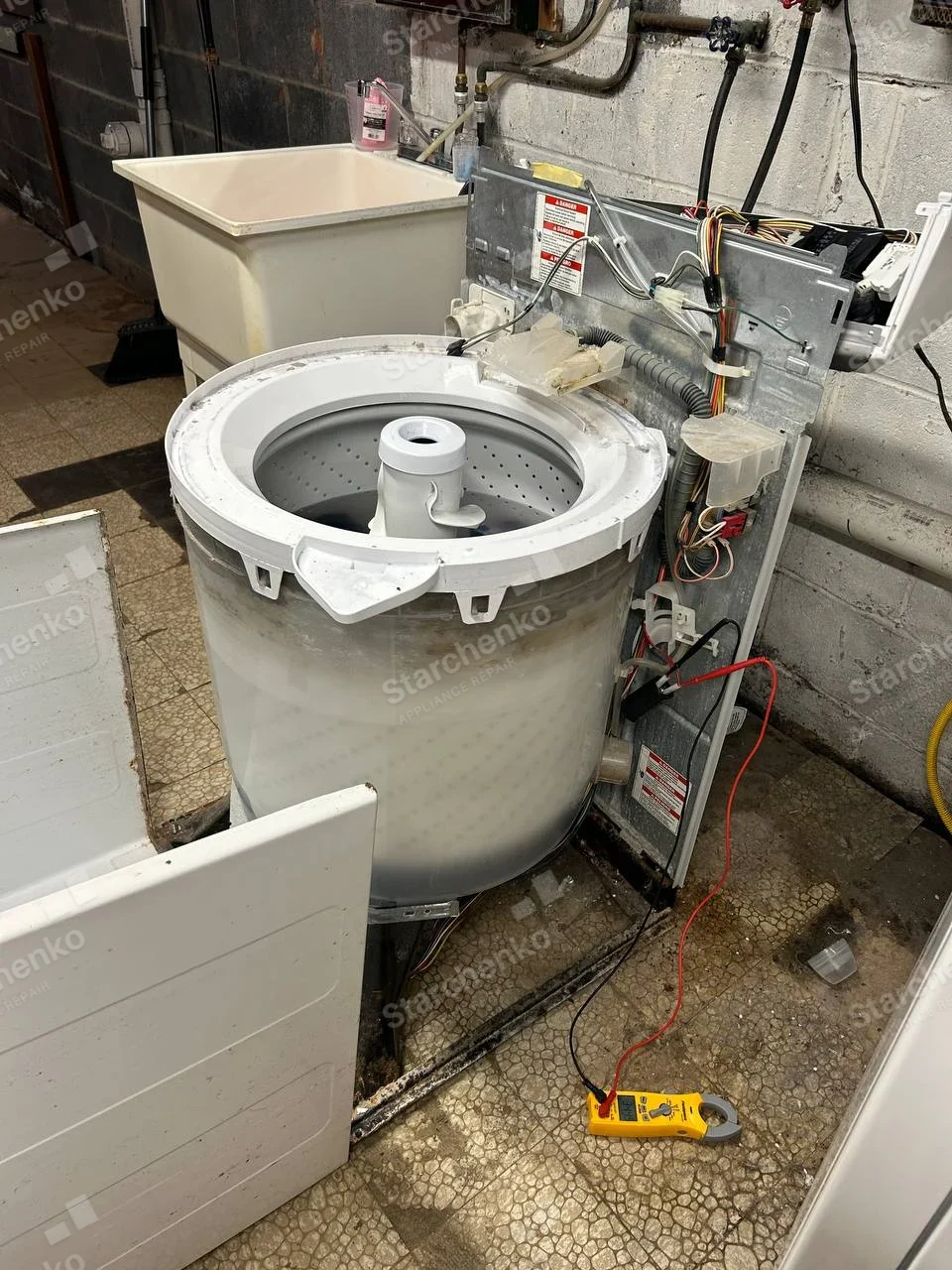 Amana repair: Top-load washer extensively disassembled with outer cabinet aside, exposed perforated basket and agitator, laundry sink adjacent.