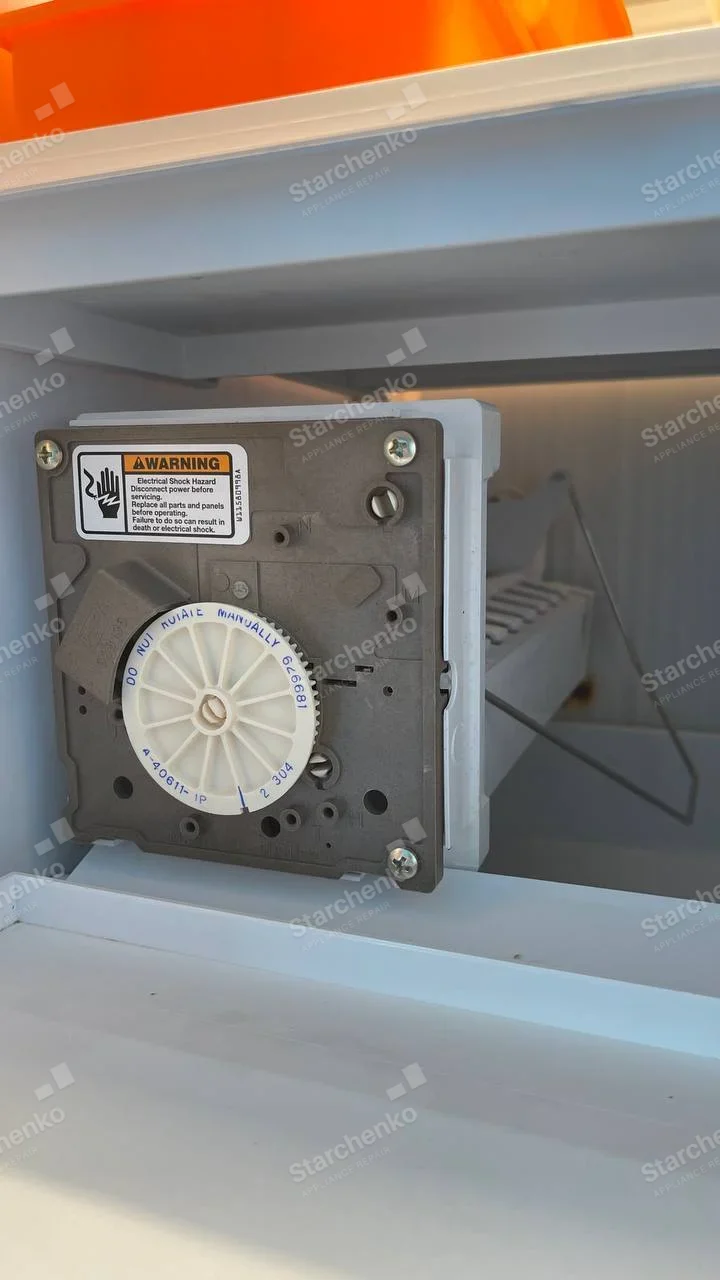 Samsung repair: Close-up ice-maker motor module and ice tray mounted inside refrigerator freezer compartment, no appliance logo.