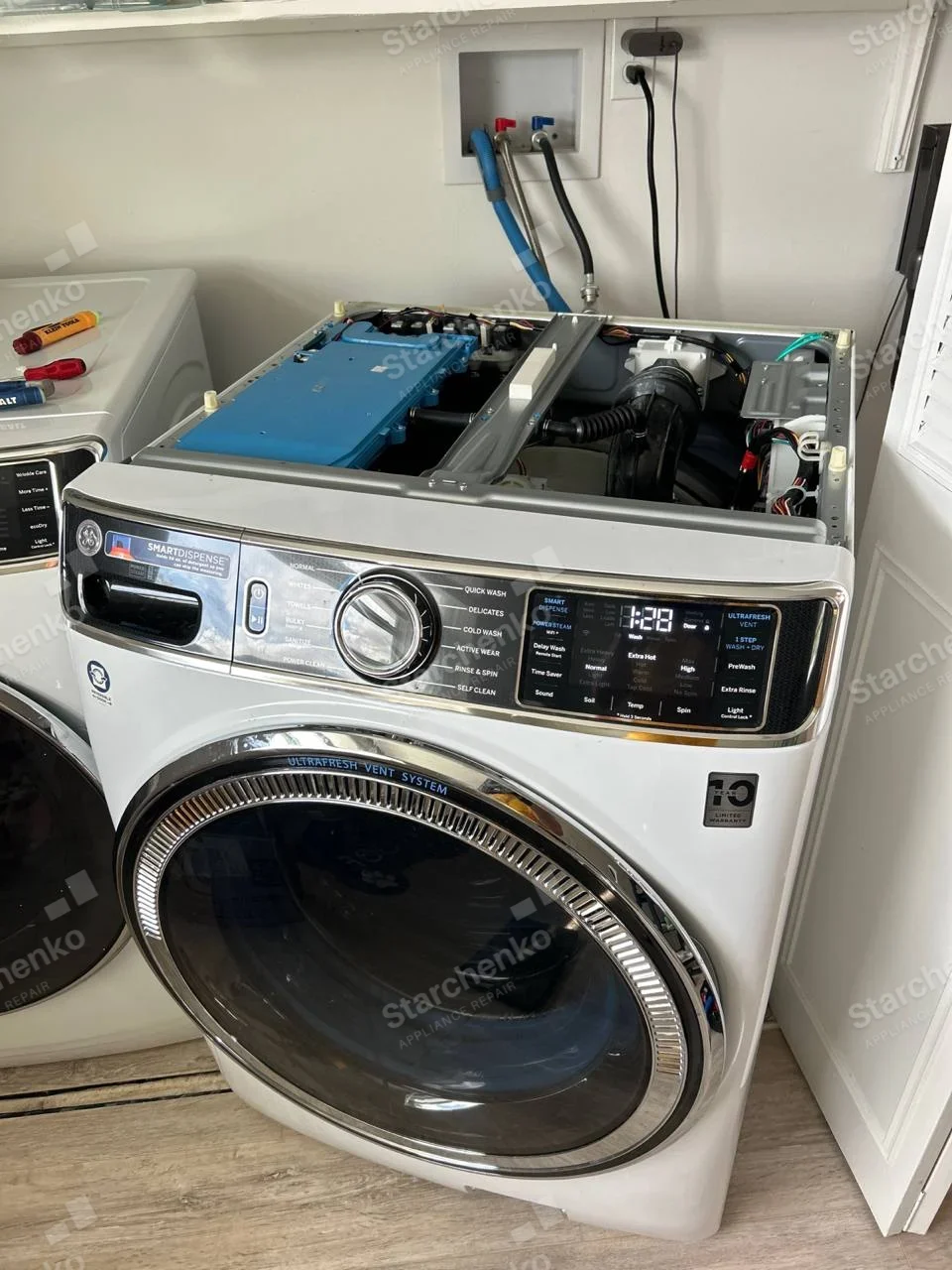 GE logo at upper left and UltraFresh Vent System around front-load door, top cover off showing blue dispenser assembly and hoses.