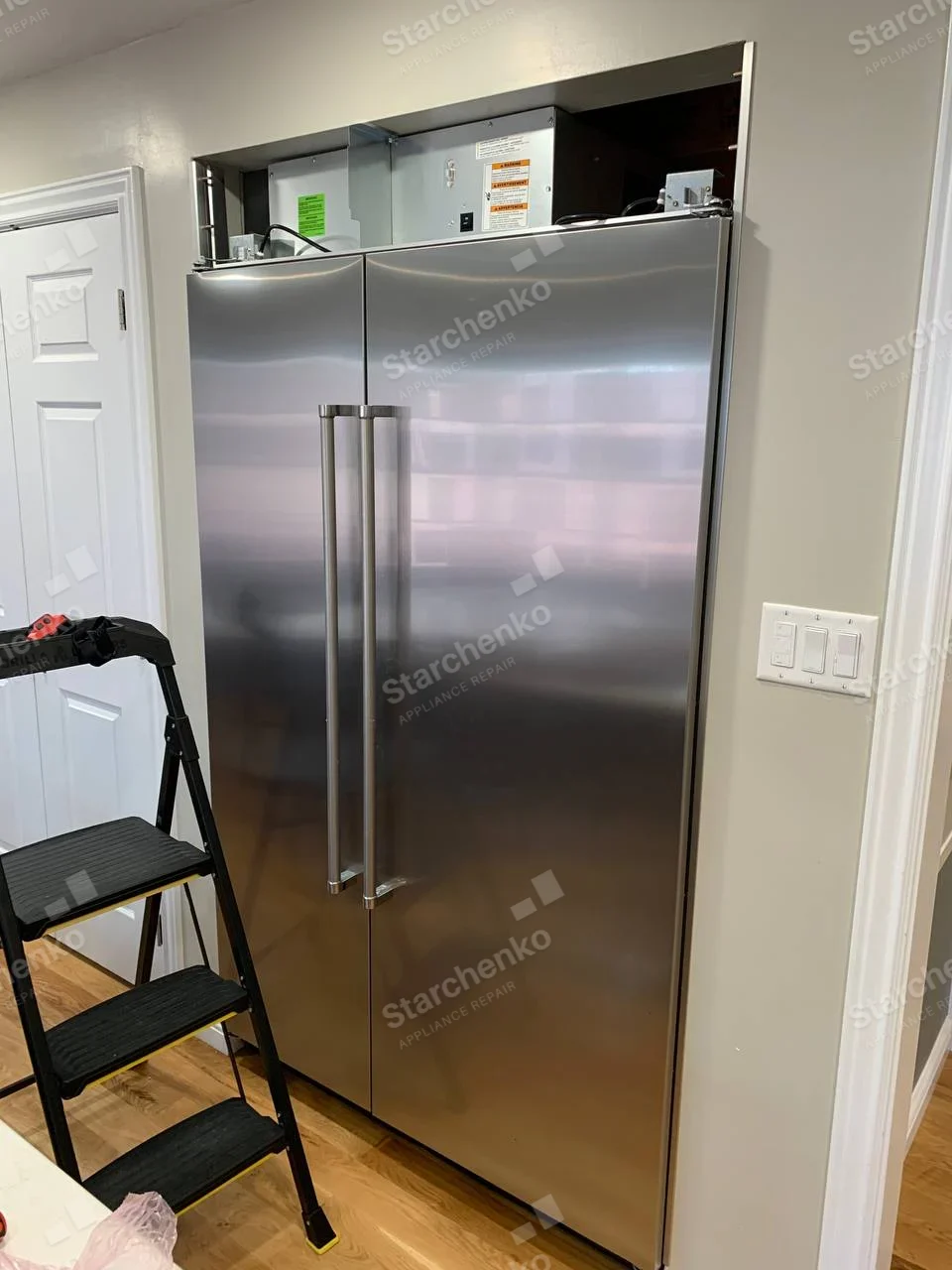 GE Monogram repair: Stainless built-in side-by-side refrigerator with open upper mechanical compartment.