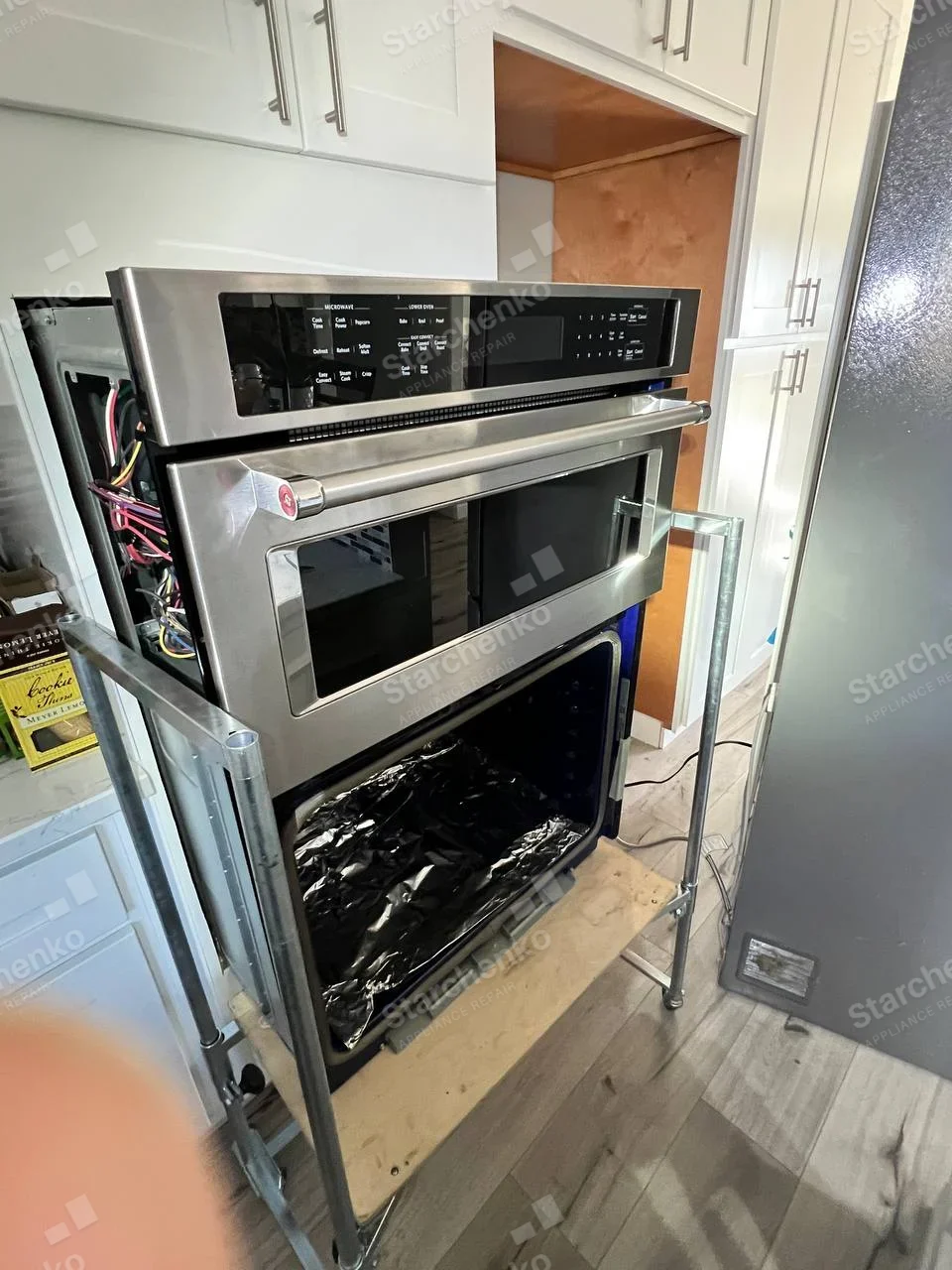 Samsung repair: Built-in microwave-and-oven stack removed on cart, lower oven door removed revealing cavity, side wiring exposed.
