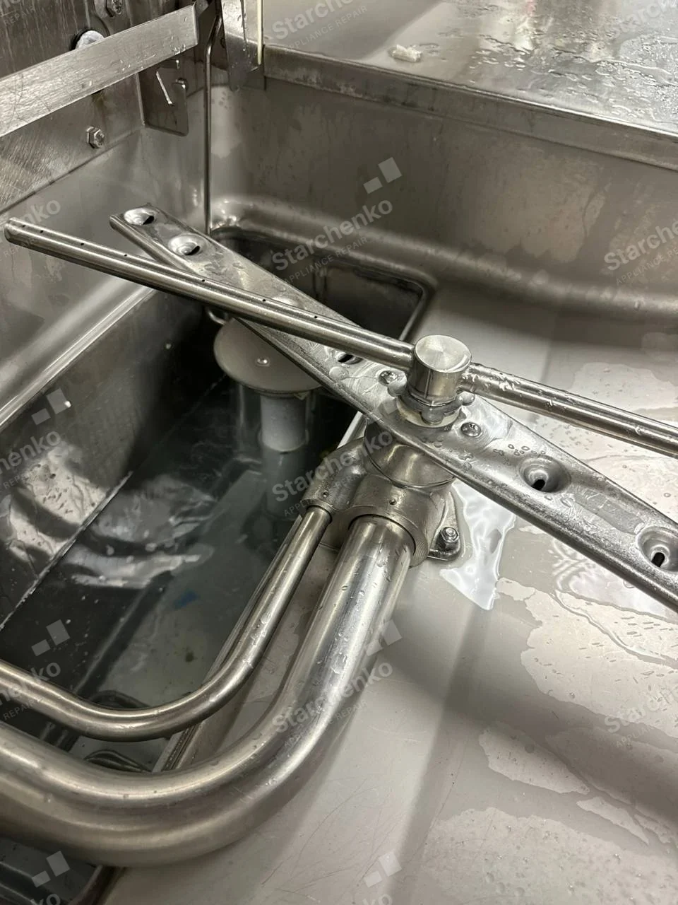 Champion repair: Inside commercial dishwasher wash tank with metal spray arm, piping and standing water.