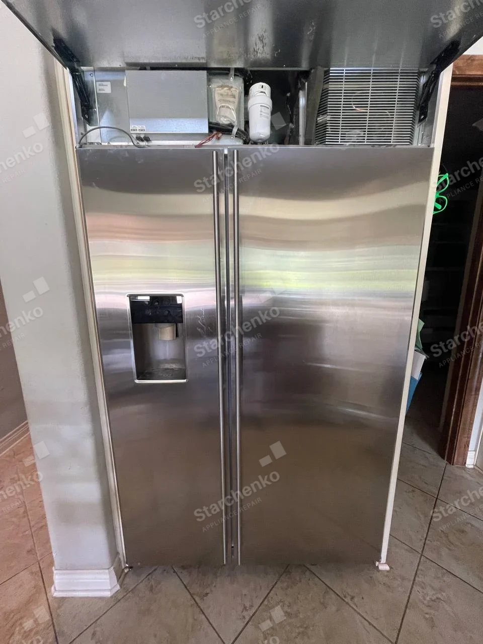 KitchenAid repair: Stainless side-by-side refrigerator with top service panel raised and upper components exposed.