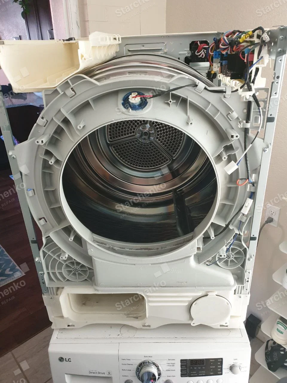 Electrolux repair: Upper dryer in laundry stack stripped of its front panel, revealing metal drum, lint duct and wiring, LG logo only on lower washer.