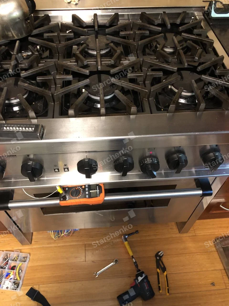 Dacor repair: Wide six-burner gas range with lower front panel removed and test meter on oven handle.