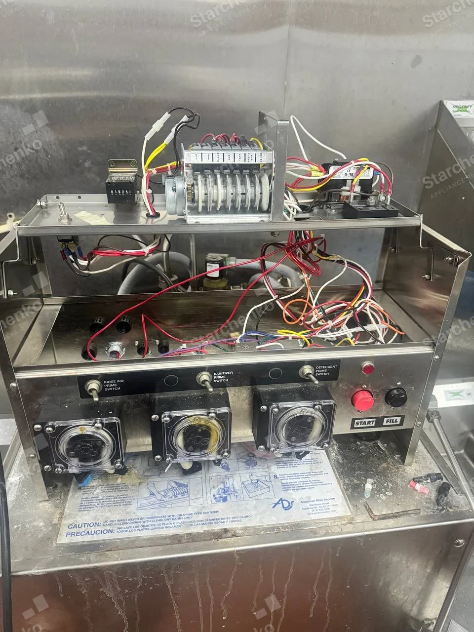 Jackson repair: Commercial dishwashing equipment open at chemical dispenser and timer controls, ADS marks appear on dispenser and instructions, not confirmed on primary machine.