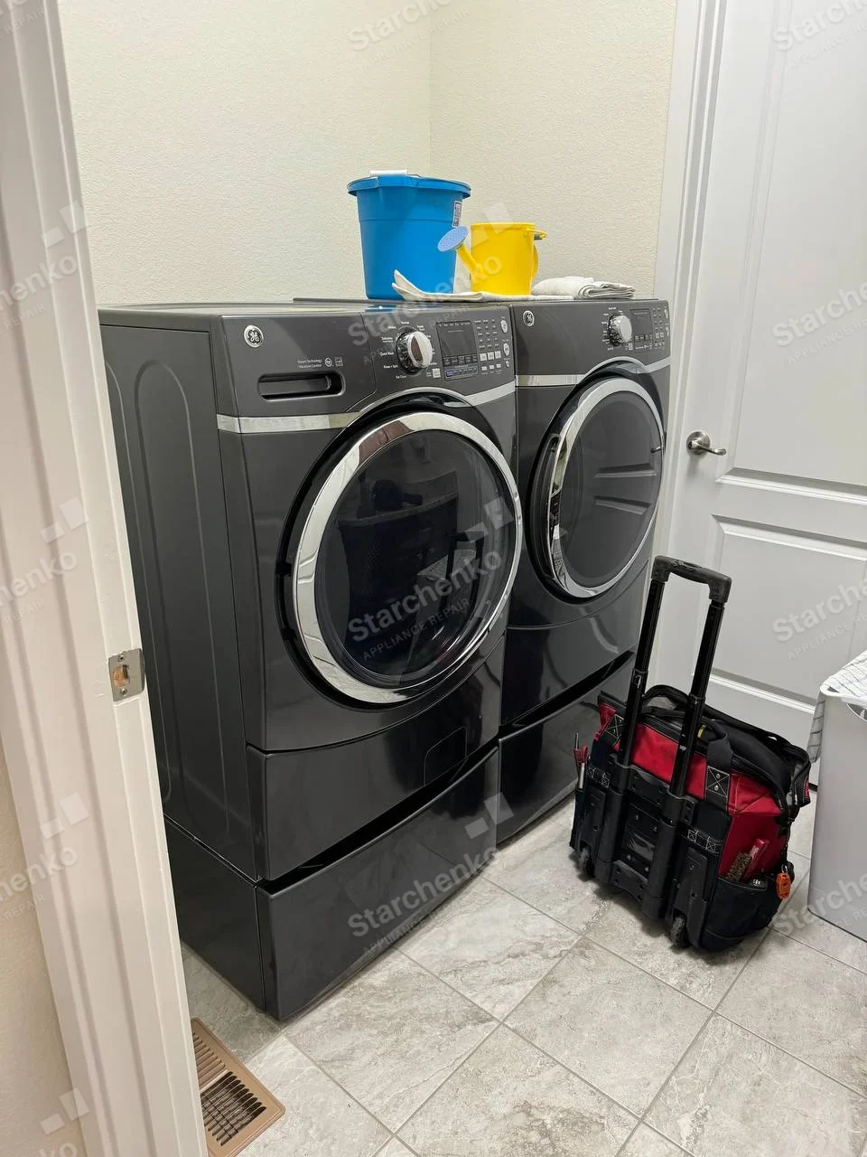 GE logo on left front-load washer's top-left panel and separate matching dryer at right, both closed, tool bag nearby.