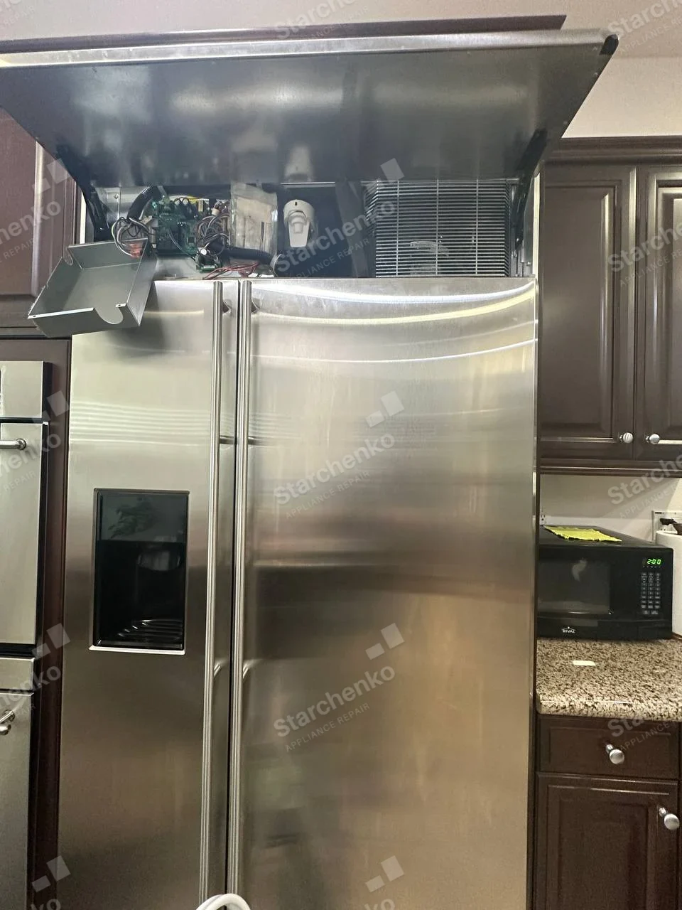 GE Monogram repair: Built-in stainless side-by-side refrigerator with dispenser, upper service cover raised exposing cooling coil and control wiring, countertop microwave separate.