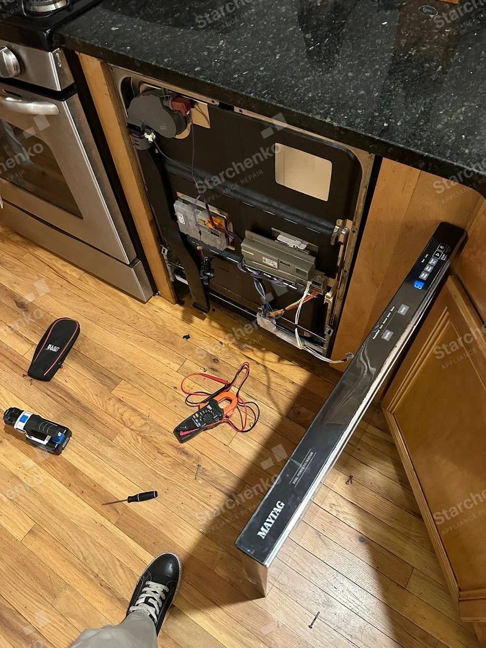 Maytag repair: Built-in dishwasher with front door and control fascia removed, exposing electronics and wiring, detached branded fascia in foreground.