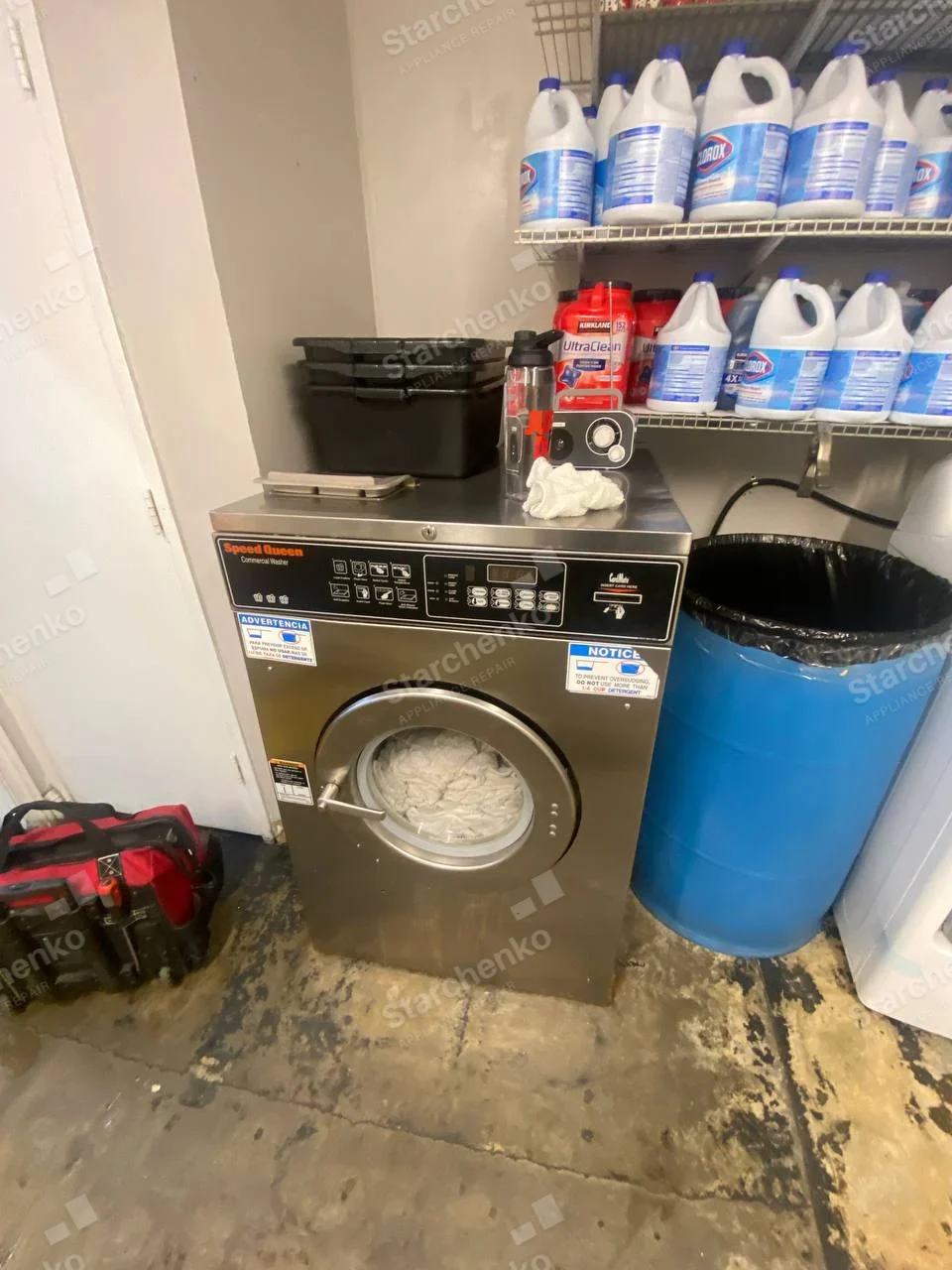 Speed Queen Commercial Washer printed above controls, front-load stainless unit in supply room.