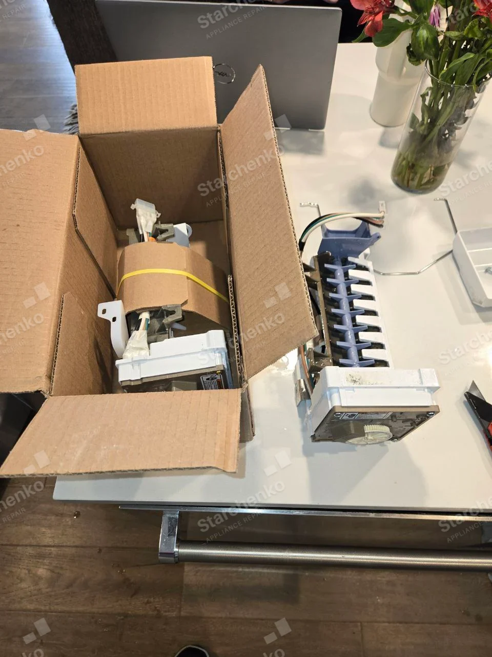Samsung repair: Detached refrigerator/freezer ice-maker assemblies on tabletop with cube ejector comb and molded ice tray visible, appliance host not shown.