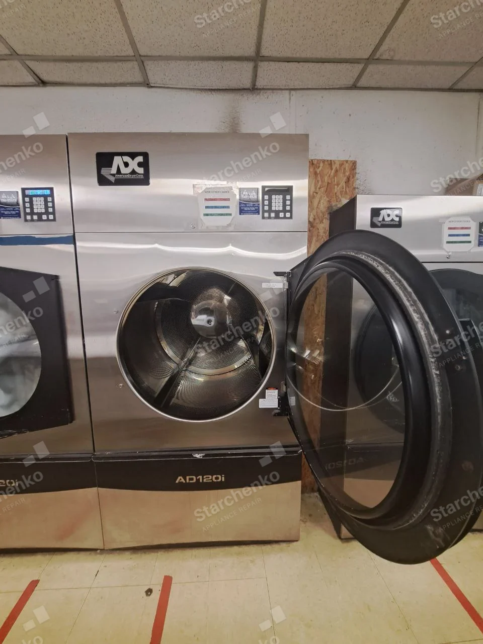 American Dryer (ADC) stainless industrial tumble dryer with large open door and perforated drum visible.