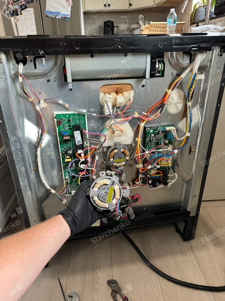 Amana repair: Exposed rear of a cooking range with two electrical boards and oven fan motors, refrigerator in background is not the repair target.