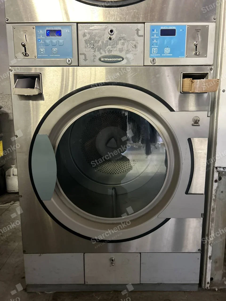 Wascomat badge centered between two controls on commercial laundry dryer, perforated drum and lint opening visible.
