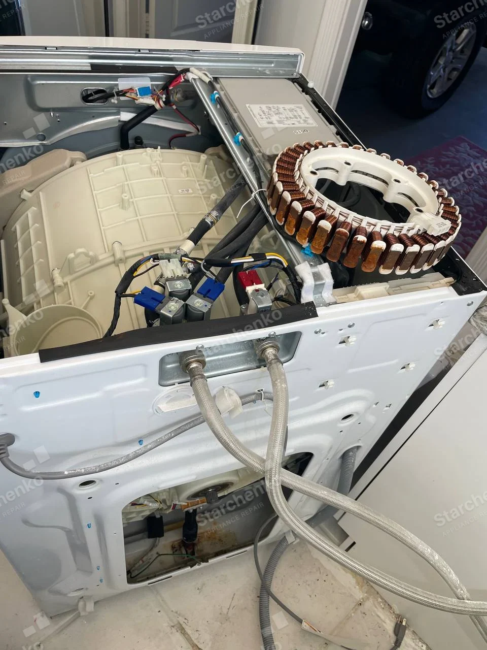 Electrolux repair: Rear of front-load washer with top and rear access removed, detached circular stator motor assembly atop tub, inlet hoses visible.