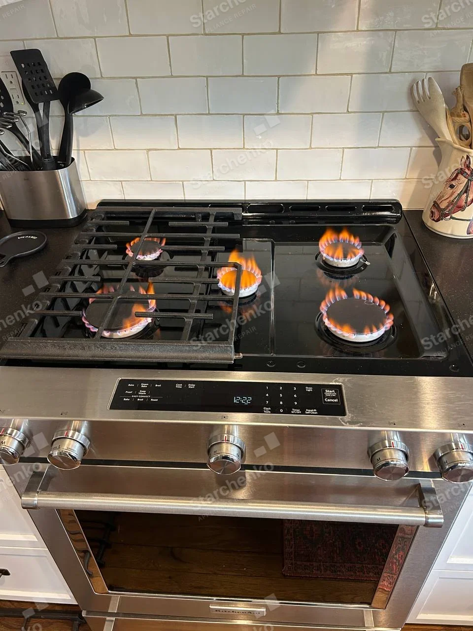 KitchenAid badge beneath oven window of gas range, all five burner flames visible in test.